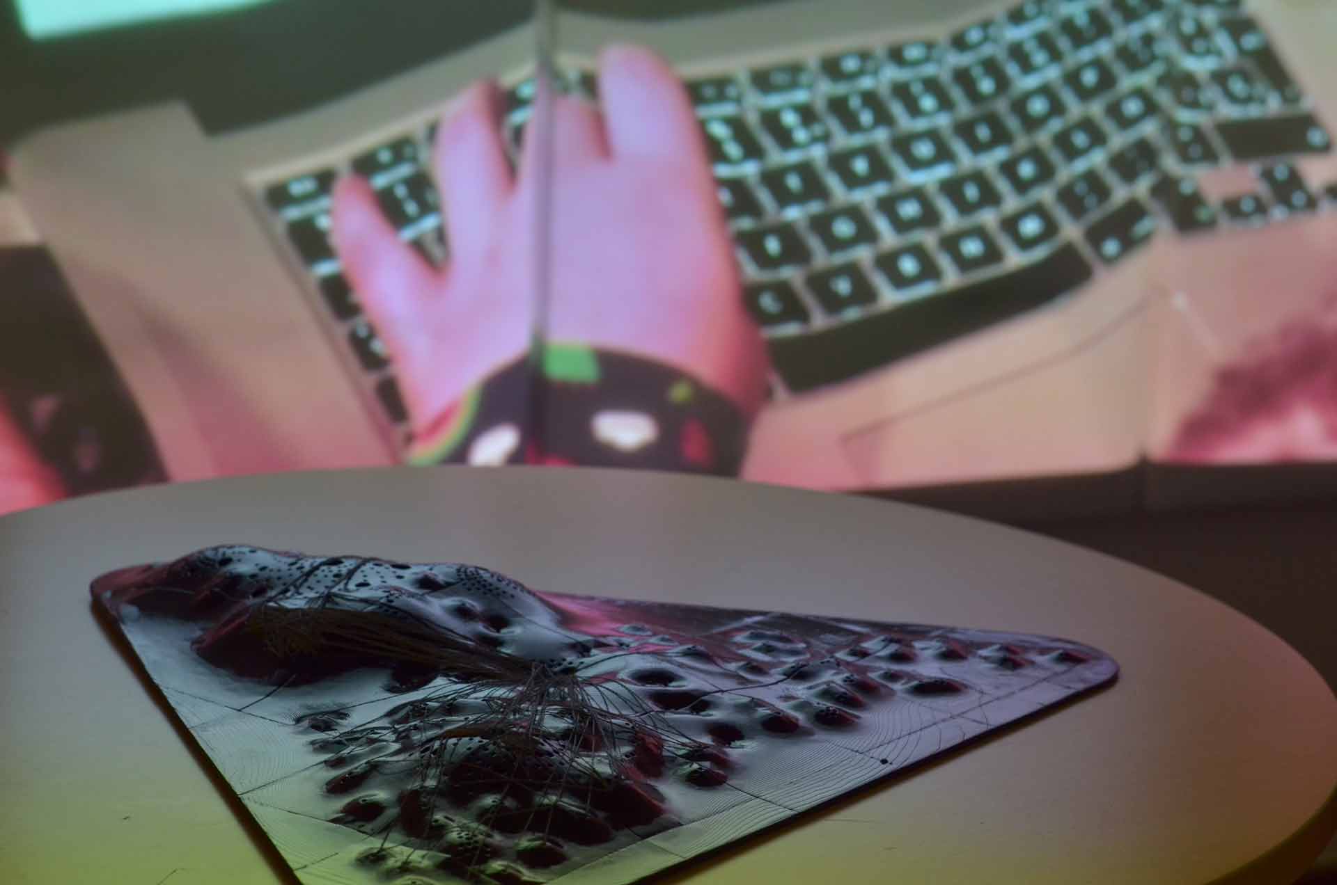In the foreground is the artwork, a topographic object with conductive threads, in the background is a projection on a wall of a hand touching a laptop keyboard.