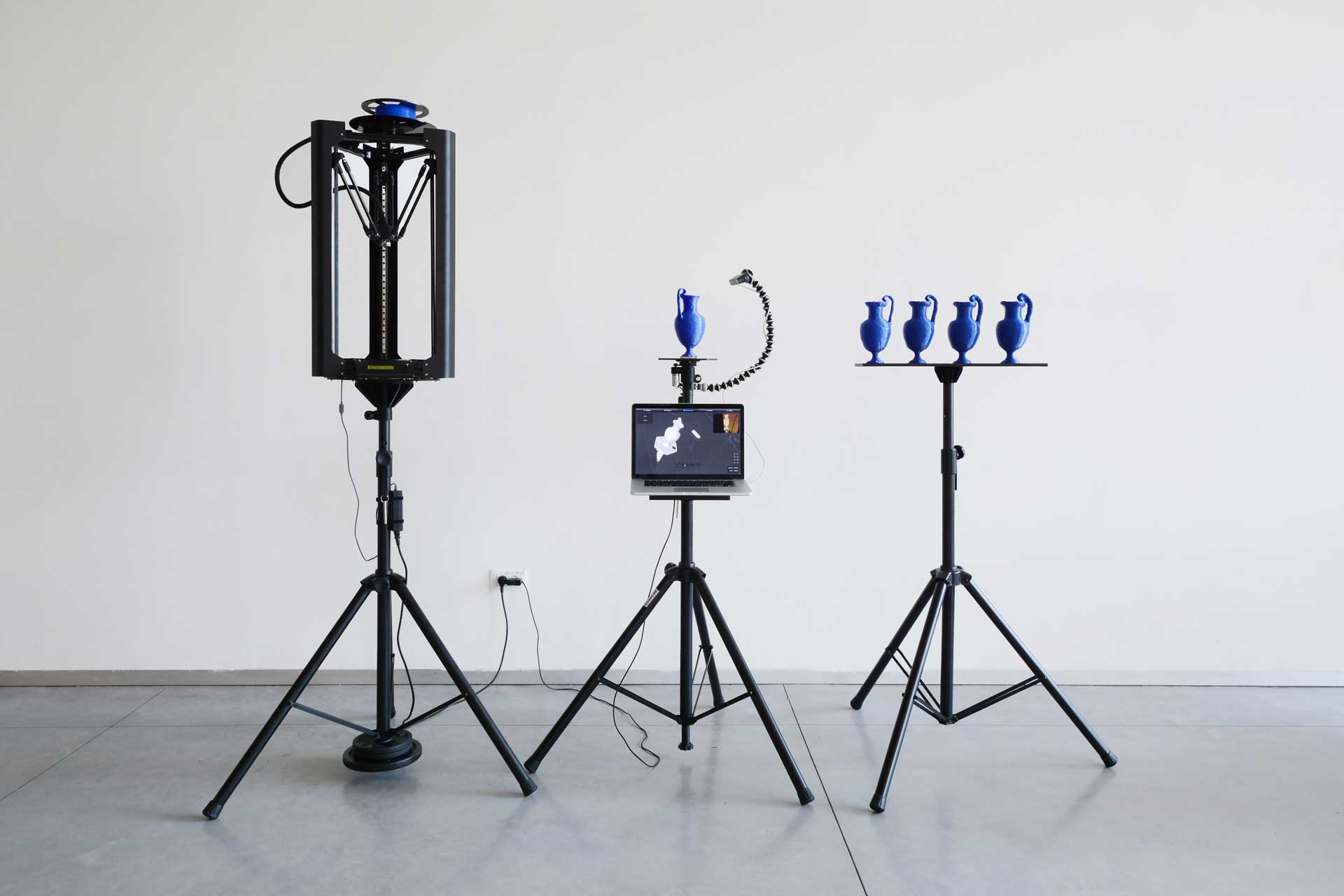 Three tripods supporting a 3D printer with blue filament; a 3D-scanning setup with a laptop, a 3D-printed blue jug, and a 3D scanner; a platform presenting four 3D-printed blue jugs.